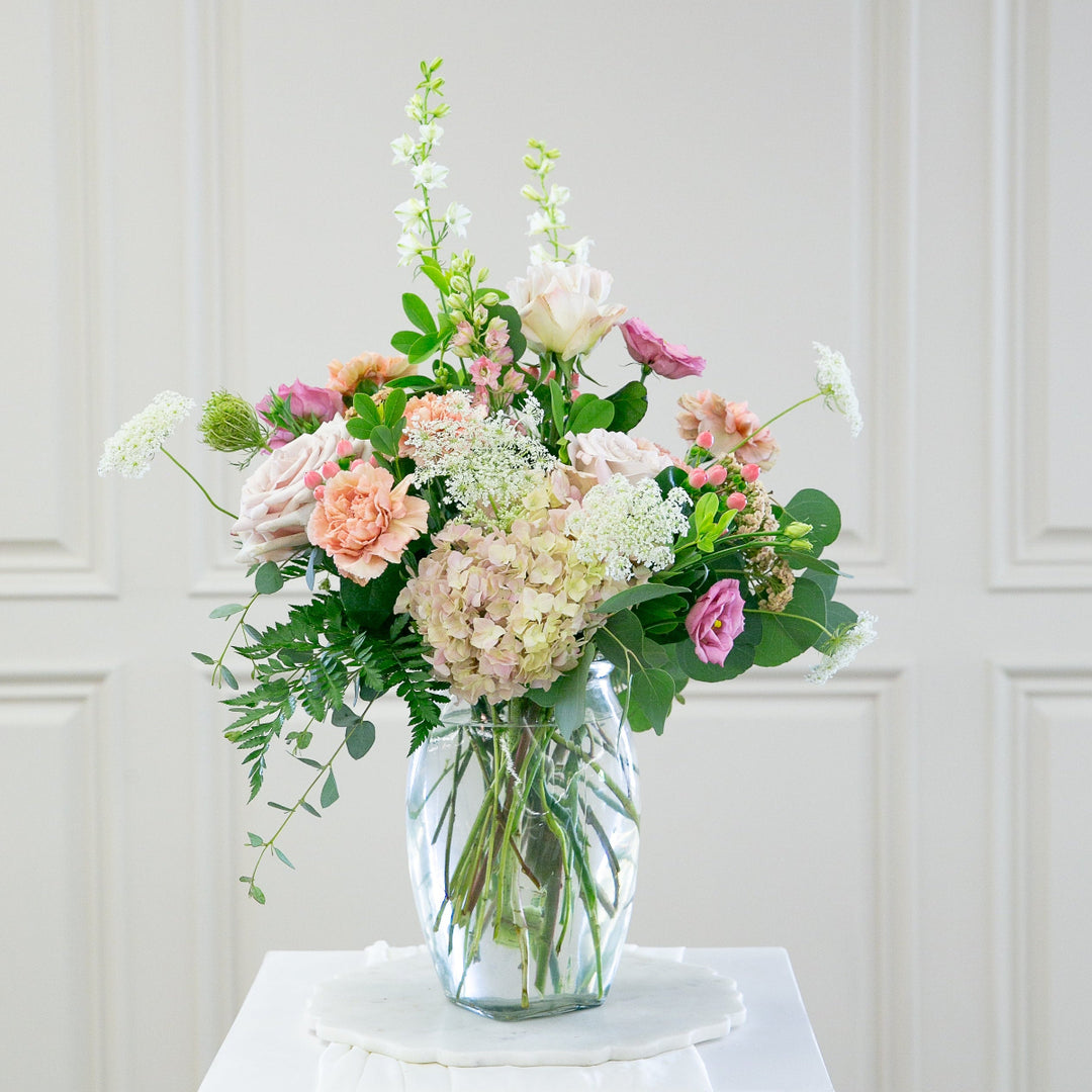 Bouquet of flowers in a clear vase on a white pedestal against a white paneled wall.