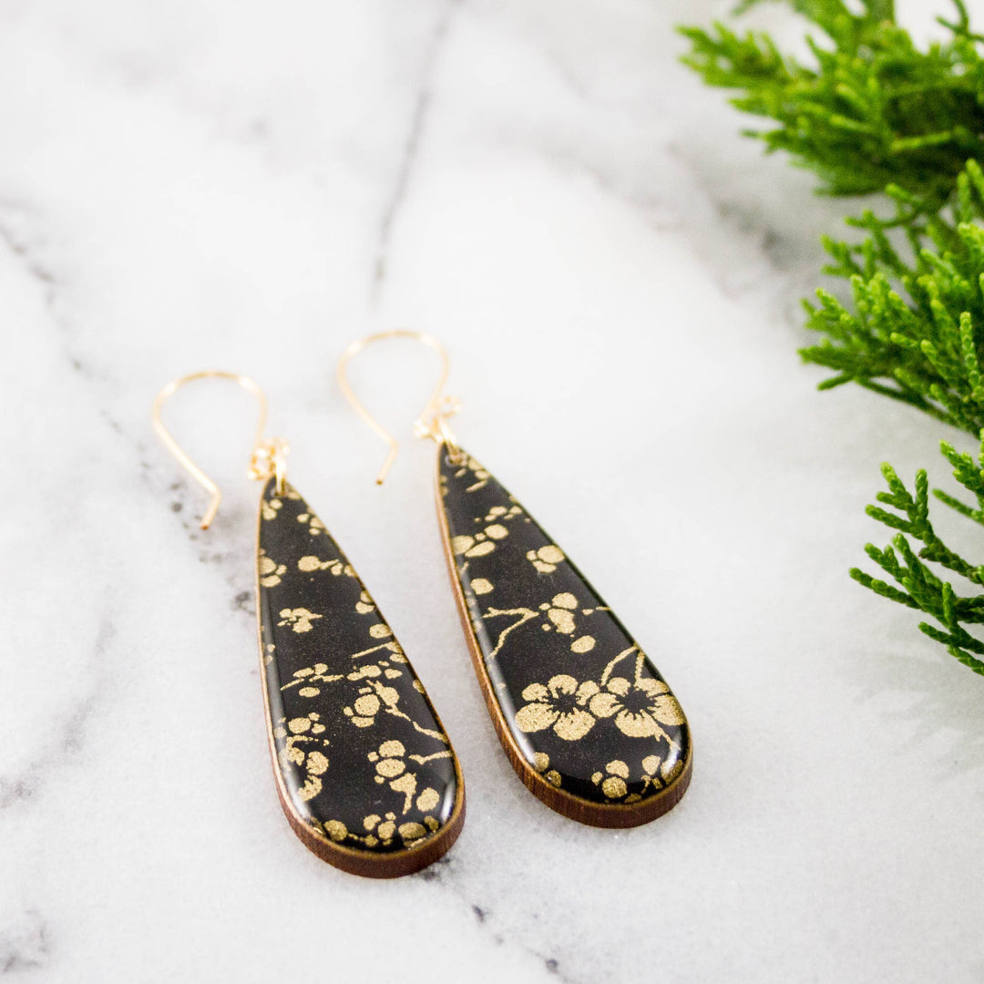 Teardrop-shaped earrings with gold floral pattern on black background, placed on a marble surface with greenery.