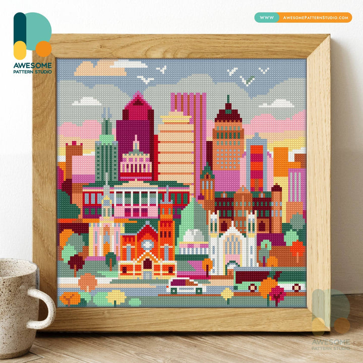 Framed cross-stitch cityscape with colorful buildings on a wooden floor.