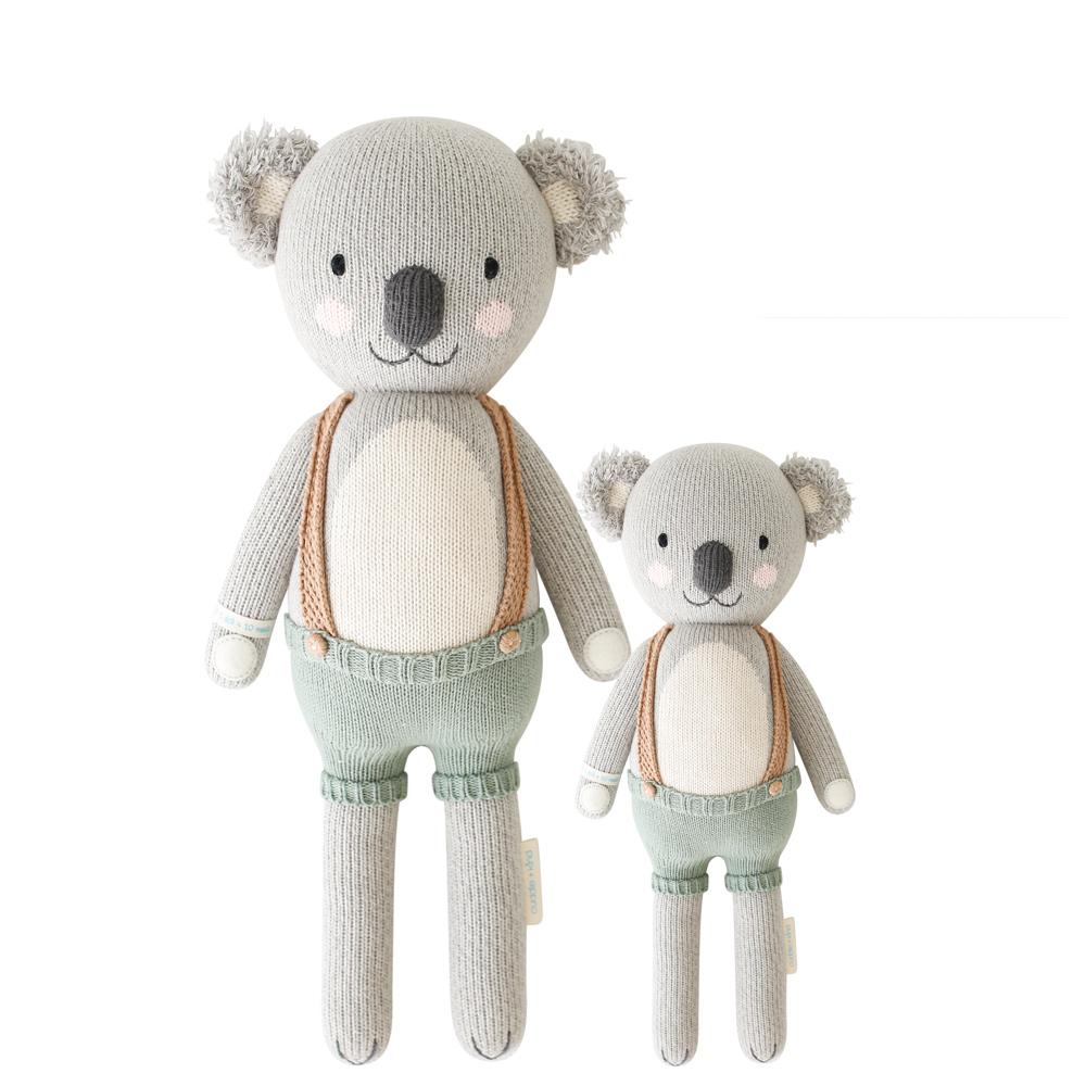 Quinn the Koala | Plush Toy | Cuddle + Kind | A gray knitted koala plush toy with light blue suspenders that have a light brown strap. Two sizes pictured, 20" and 13",