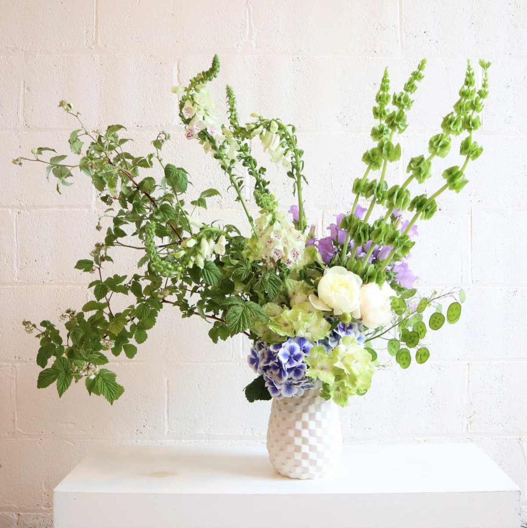 Large floral arrangement with mostly green florals with pops of blue and purple against a white background.