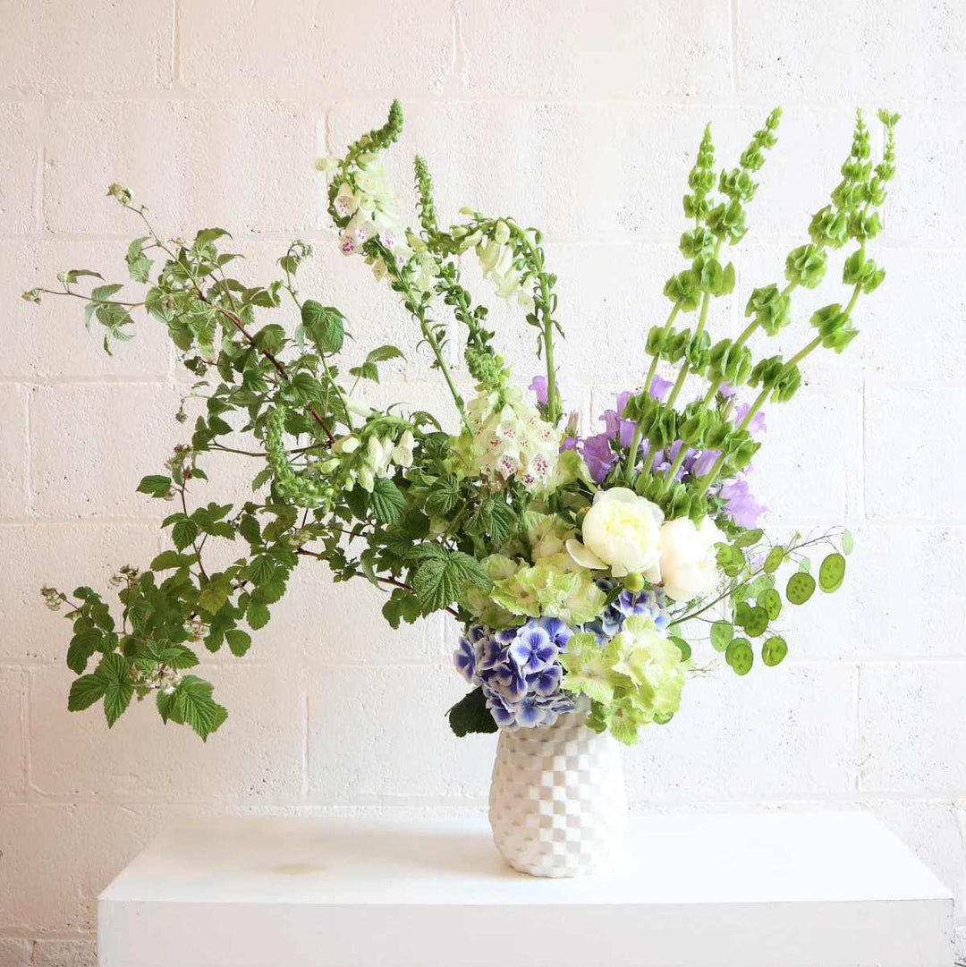 Large floral arrangement with mostly green florals with pops of blue and purple against a white background.