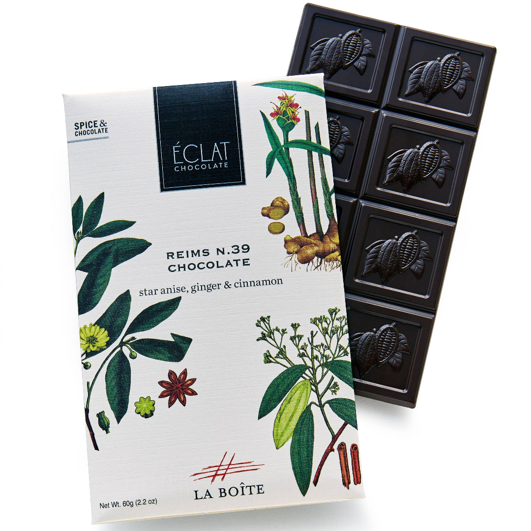 A bar of La Boite Chocolat Reims N.39 chocolate with botanical illustrations on the white wrapper, shown next to a dark chocolate bar segment on a white background.