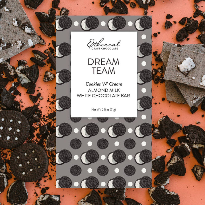 Ethereal Craft Chocolate Dream Team Cookies 'N' Cream Almond Milk White Chocolate Bar packaging surrounded by Oreo cookies on a pink background.