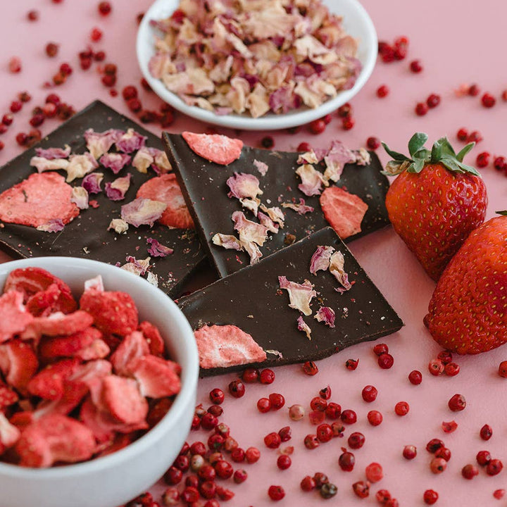 Chocolate bars with strawberry pieces on a pink surface with strawberries and a bowl of strawberry pieces.