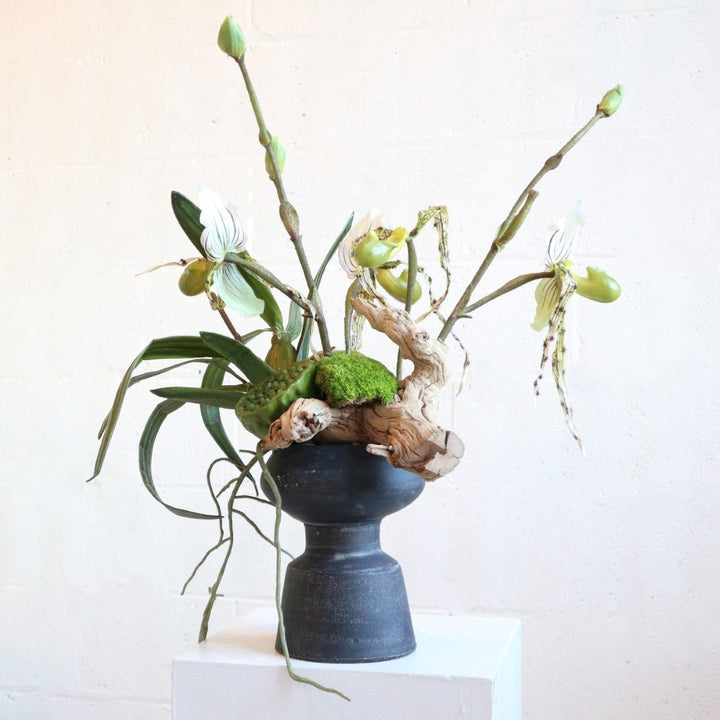 Faux flower arrangement with grapewood, lotus pods, lady slipper orchids, moss in a black vessel.