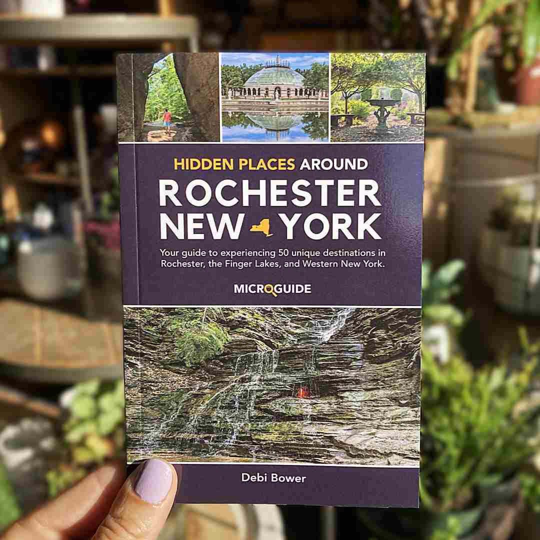 Stacy K Floral | Book titled 'Hidden Places Around Rochester New York' held by a hand with a blurred background. By Debi Bower