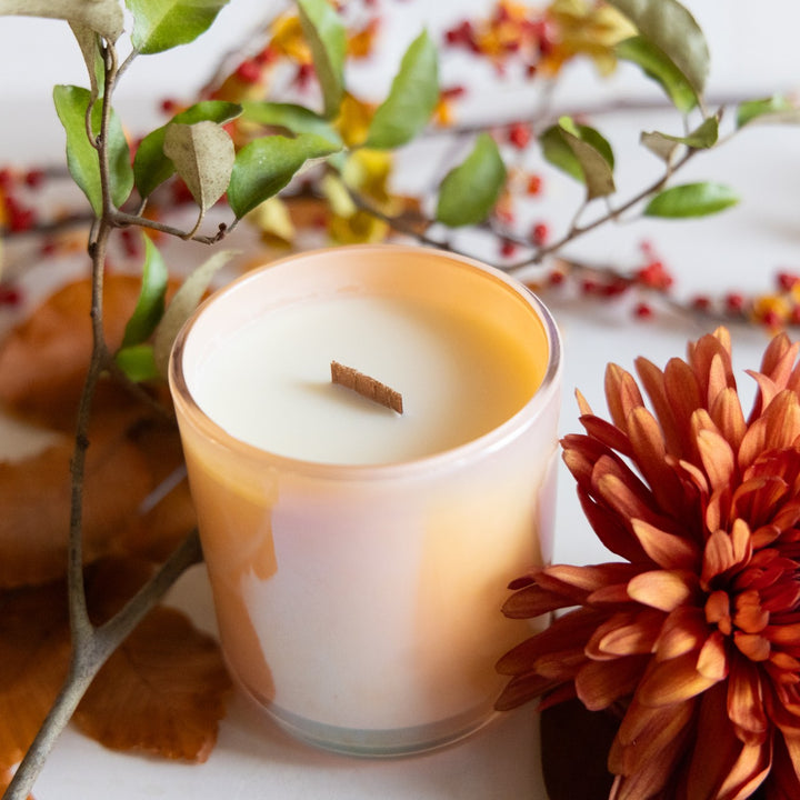 Candle in a glass holder with autumn decorations including leaves and flowers on a white surface.