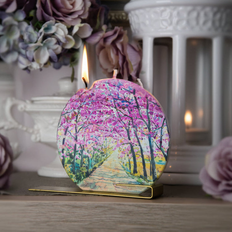 Decorative candle with a scenic design on a wooden surface with flowers and a lantern in the background.