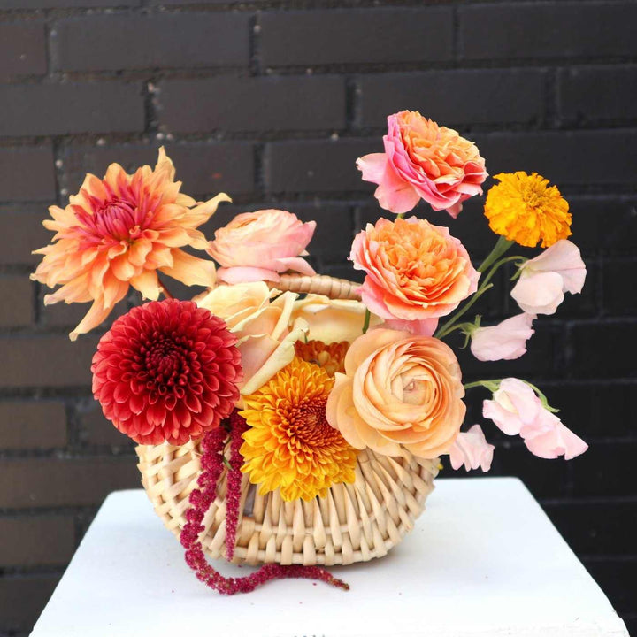 fall basket with orange dahlias, bronze mums, ranunculus, marigolds, light pink sweet pea, and peach roses on a black brick background.