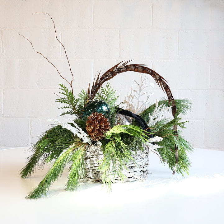Holiday Feather | A winter arrangement in a white nest basket. Contains a keepsake ornament, pinecones, feathers, flocked branching, and evergreen branching.
