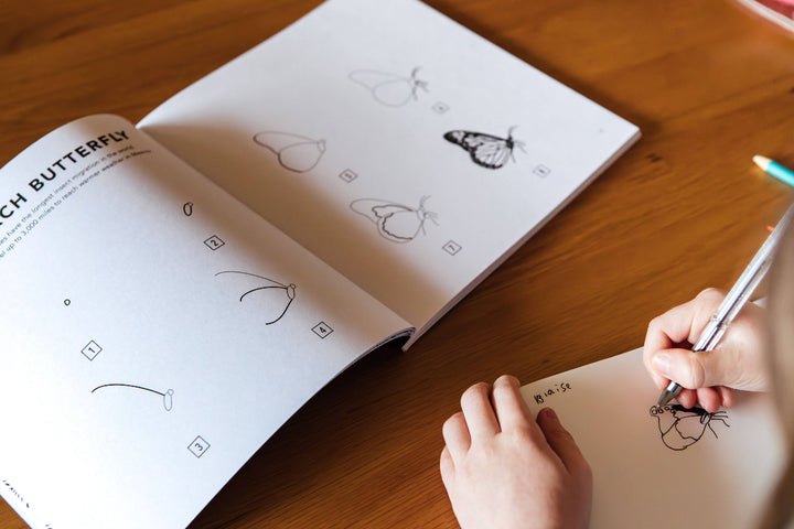 Person drawing a butterfly in a sketchbook on a wooden table