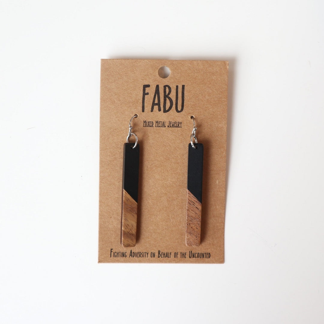 Fabu Earrings | Black stained and natural wood dangling earrings. On a brown paper backing. Reads "Fabu, mixed metal jewelry, Fighting Adversity on Behalf of the Uncounted".