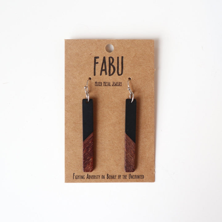 Fabu Earrings | Stained black wood and natural dark wood dangling earrings.  On a brown paper backing. Reads "Fabu, mixed metal jewelry, Fighting Adversity on Behalf of the Uncounted".