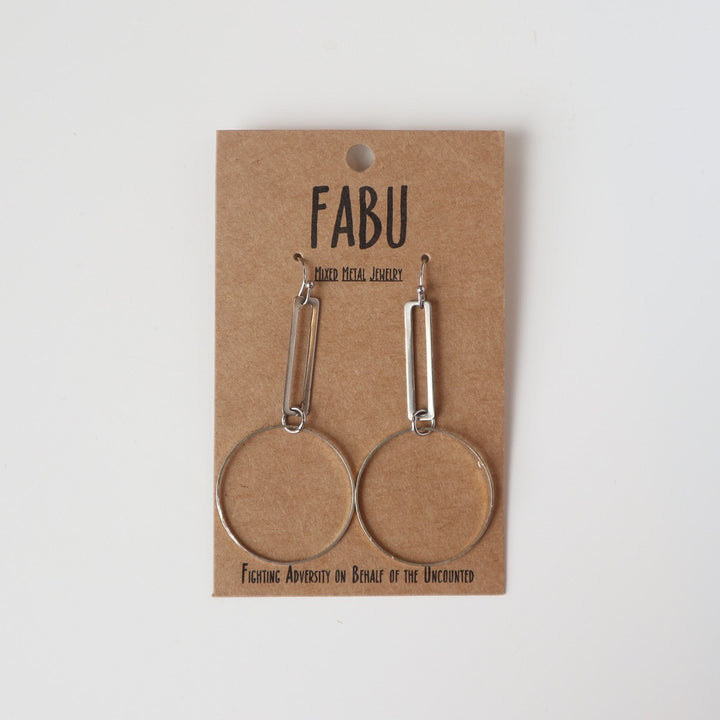 Fabu Earrings | Silver earrings with a hollow rectangle piece with a hoop hanging at the bottom.  On a brown paper backing. Reads "Fabu, mixed metal jewelry, Fighting Adversity on Behalf of the Uncounted".