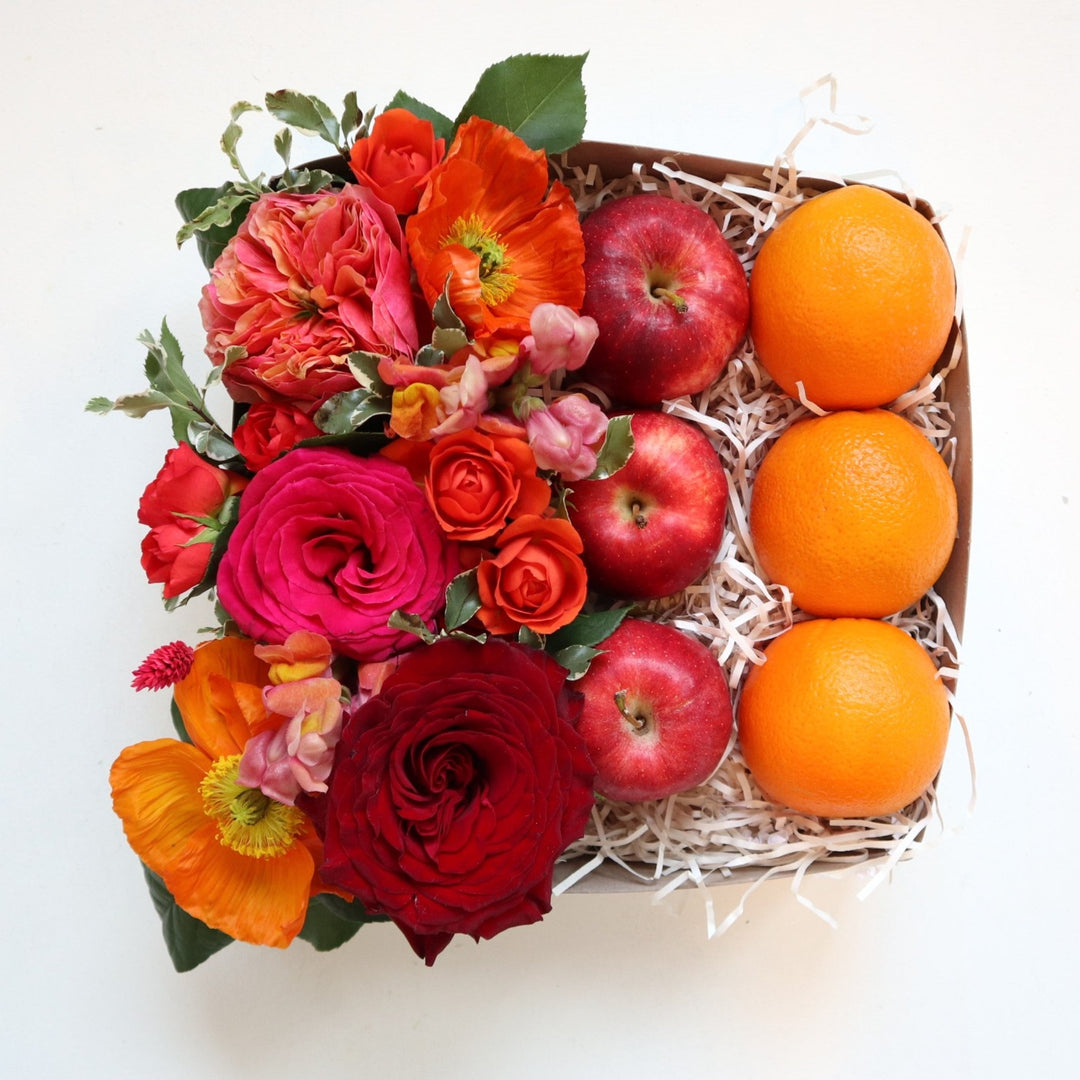 Gift box with a red rose, pink rose, orange spray rose, orange poppies and greenery alongside three apples and three oranges.