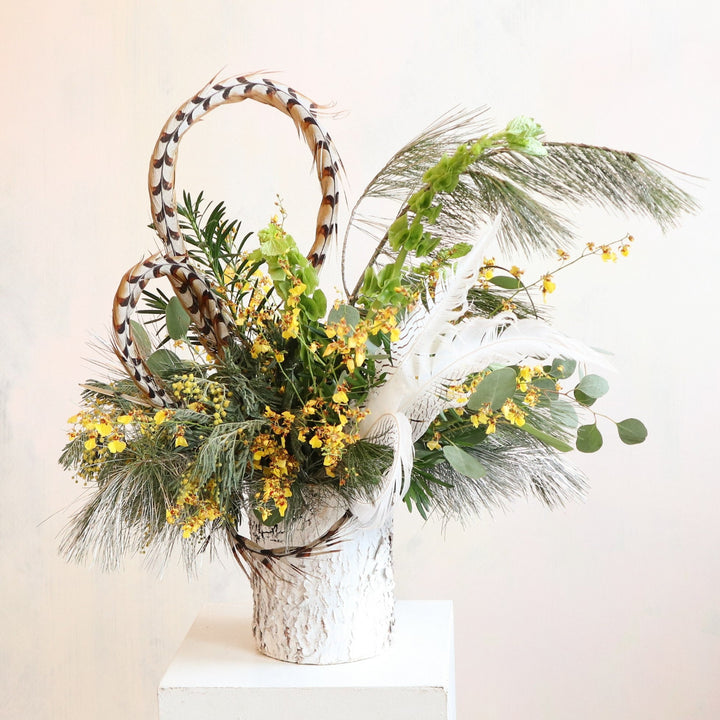 A nature-inspired floral arrangement in a rustic birch-textured vase. The design features yellow flowers, lush greenery, pheasant feathers, and white plumes, creating a dynamic and elegant composition. The feathers add height and texture, while the greenery cascades softly, evoking a woodland aesthetic.