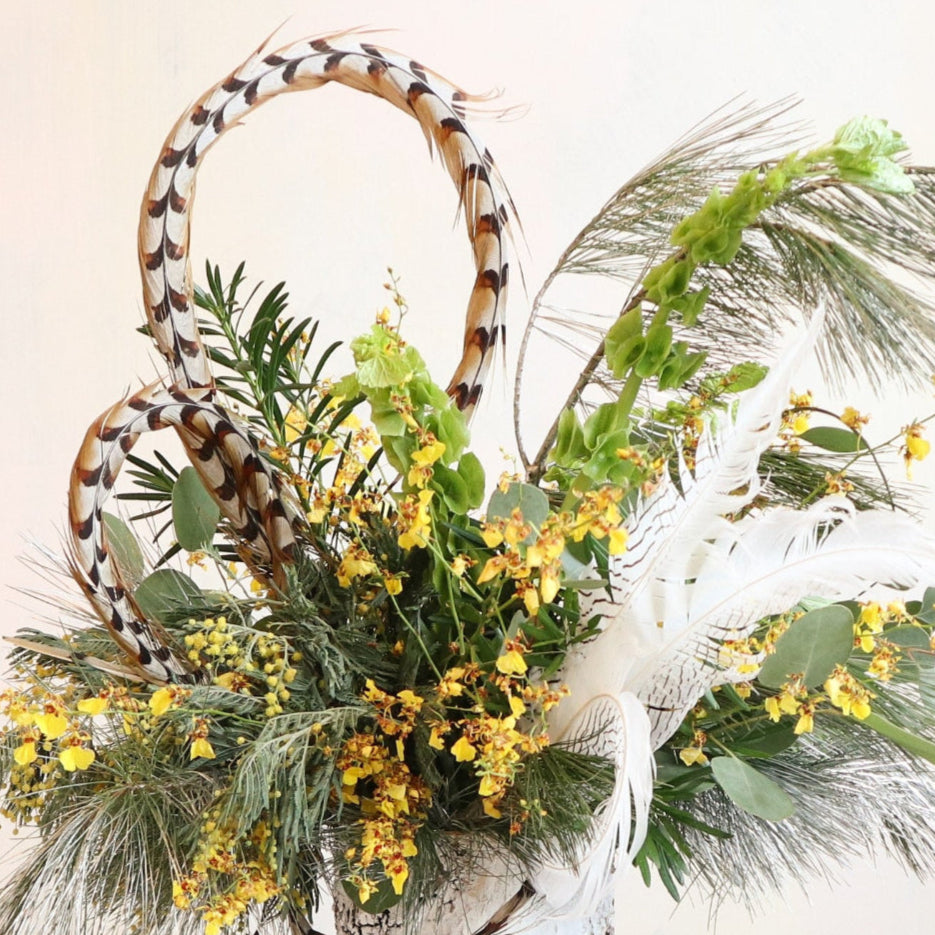 A nature-inspired floral arrangement in a rustic birch-textured vase. The design features yellow flowers, lush greenery, pheasant feathers, and white plumes, creating a dynamic and elegant composition. The feathers add height and texture, while the greenery cascades softly, evoking a woodland aesthetic.