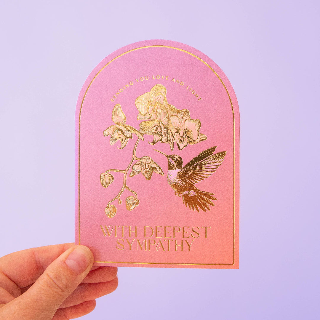 Pink card with gold hummingbird and floral design held by a hand against a light purple background