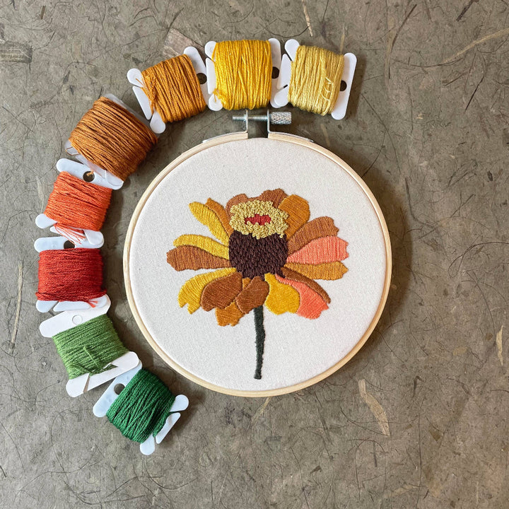 Embroidery hoop with a colorful flower design surrounded by spools of thread on a textured surface.