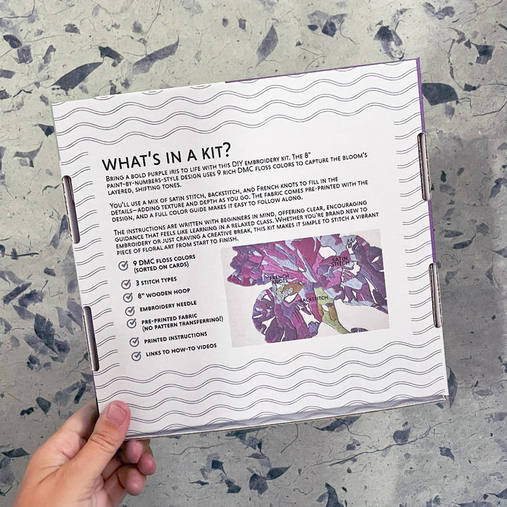 Floral embroidery kit packaging with text and a purple flower illustration on a textured surface.