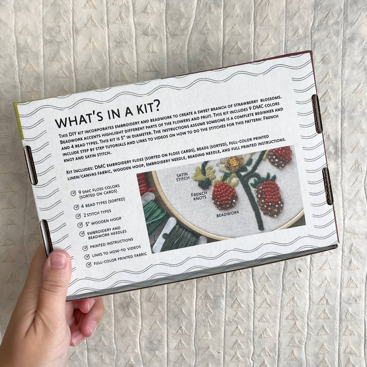 Open book showing embroidery kit description with a hand holding it against a textured wall.