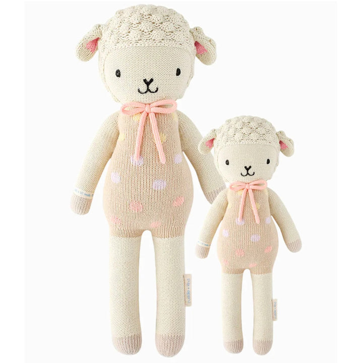 Lucy The Lamb | Cuddle + Kind | A cream colored, hand knit, lamb plushie with pink accents. The lamb is wearing a light pink/tan polka dotted outfit with a pink bow. Photo shows two sizes: 20" and 13".