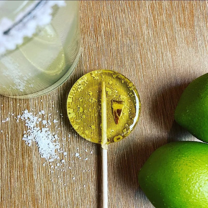 Good Lolli Lollipops | Margarita | A transparent lollipop with a lime piece and a salt rim. Photo taken with limes, salt, and a margarita in a glass.