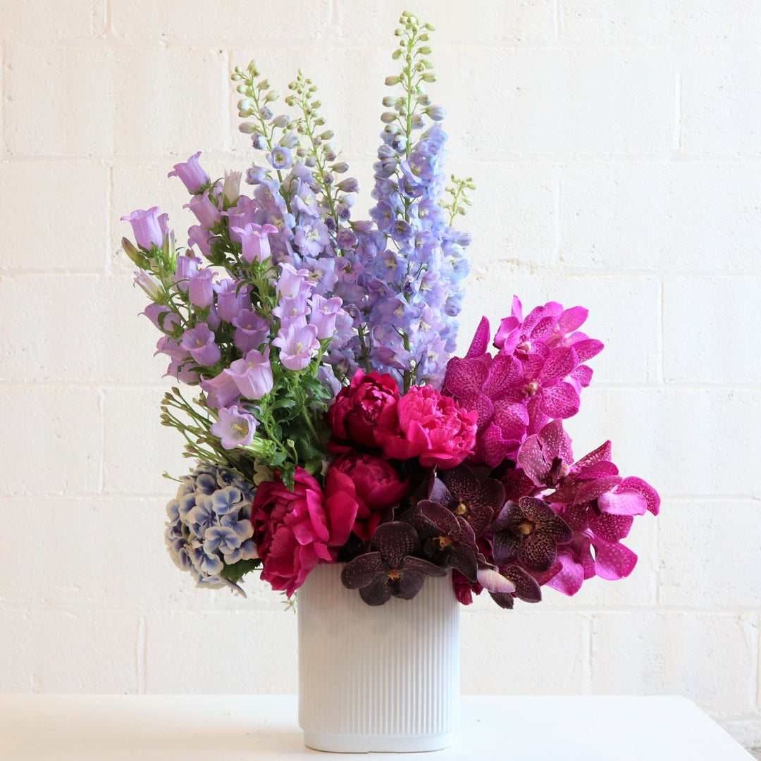 Flower arrangement with magenta and purple orchids, blue delphinium, purple campanula, blue hydrangea and pink peonies in a white vase.