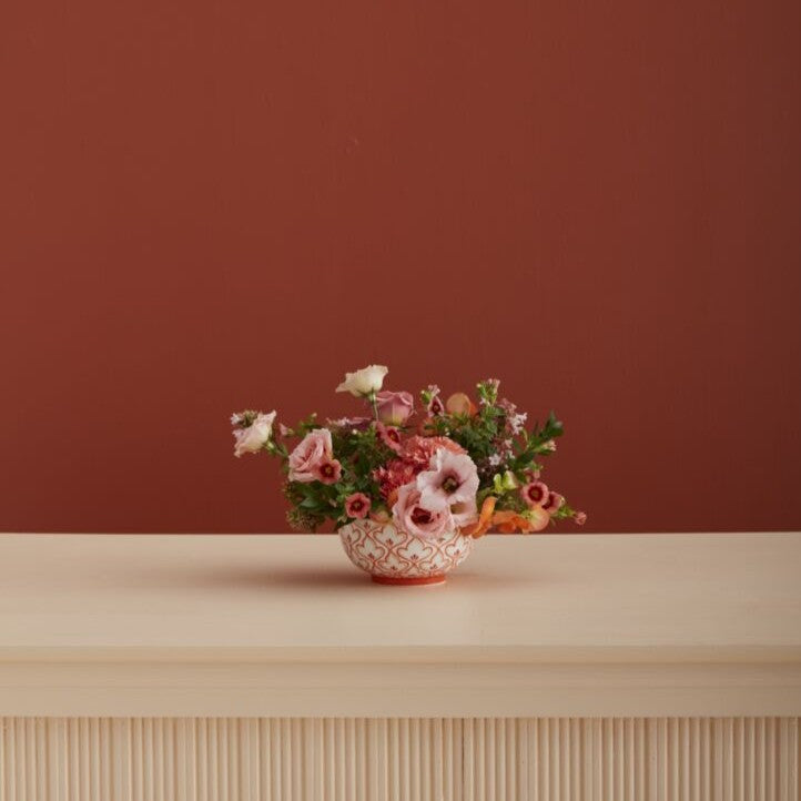 Floral arrangement in a decorative pot on a wooden surface with a red-brown background