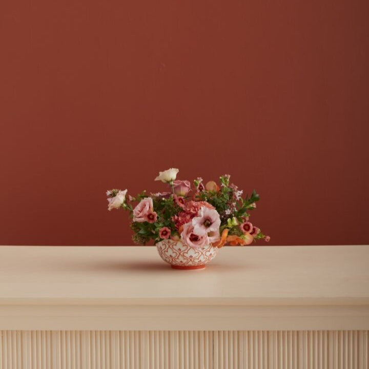 Floral arrangement in a decorative pot on a wooden surface with a red-brown background