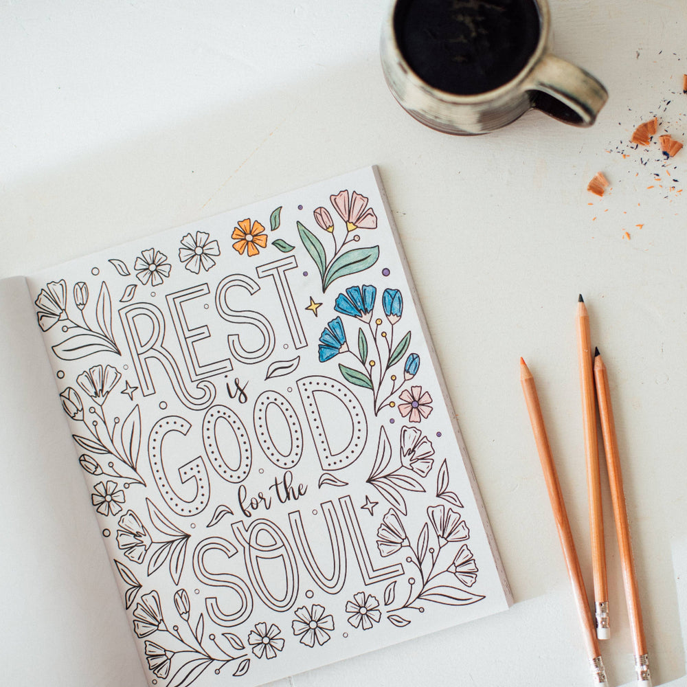 Coloring book with 'Rest is good for the soul' text, pencils, and a mug on a white surface