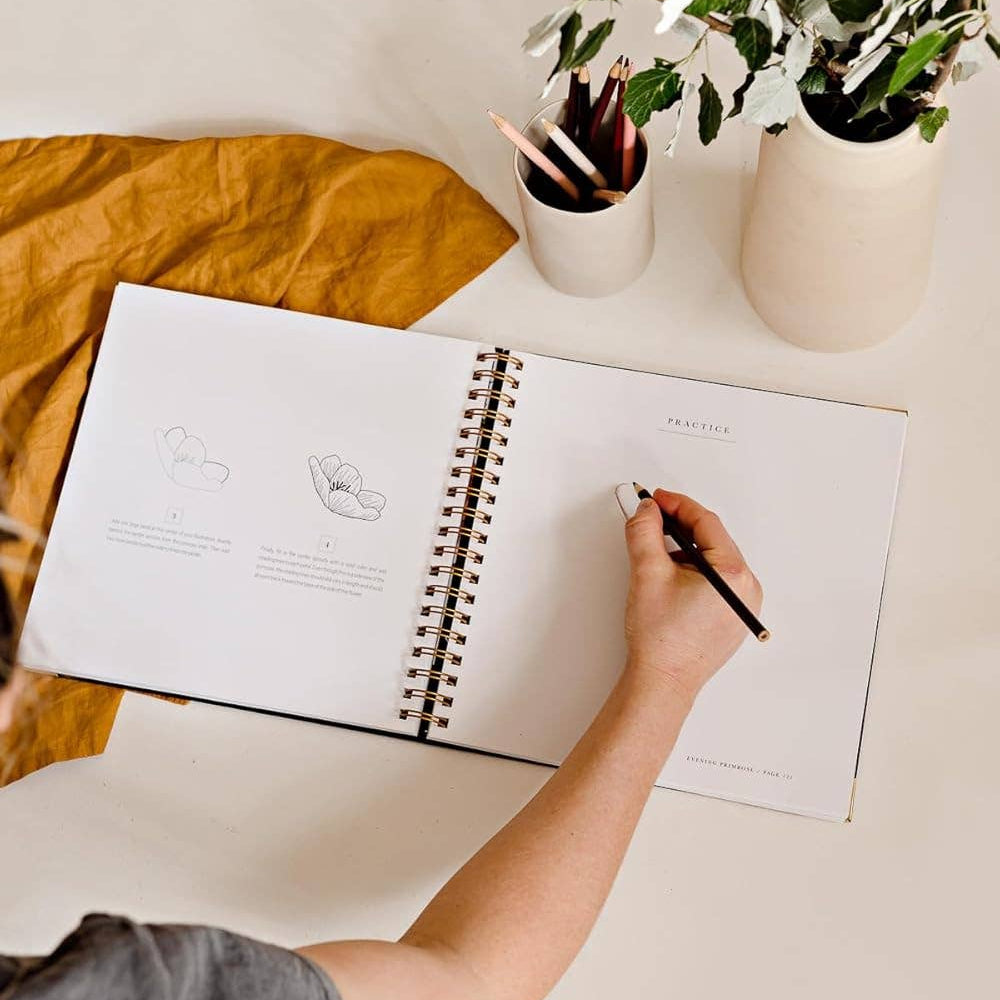Person writing in a notebook with a vase of flowers and a yellow blanket in the background