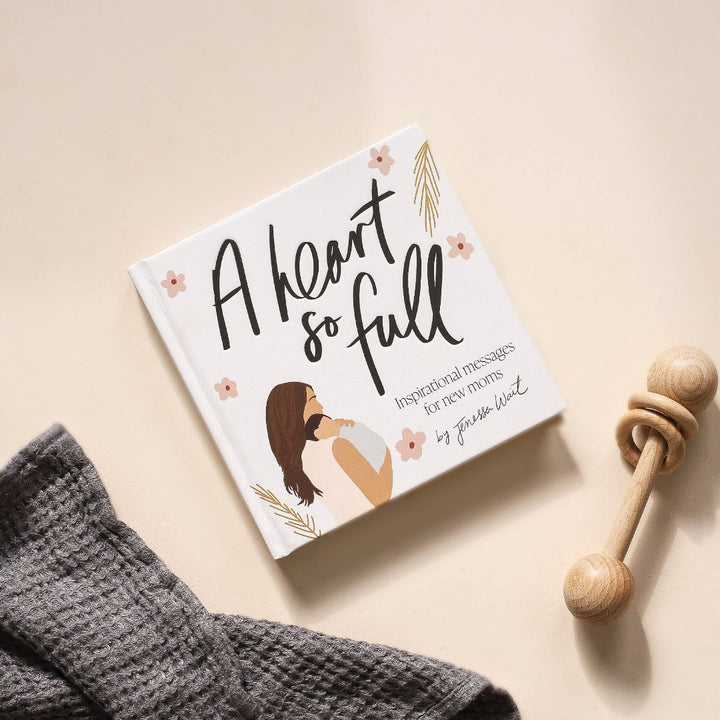 Book titled 'A Heart So Full' on a beige surface with a wooden rattle and gray blanket.