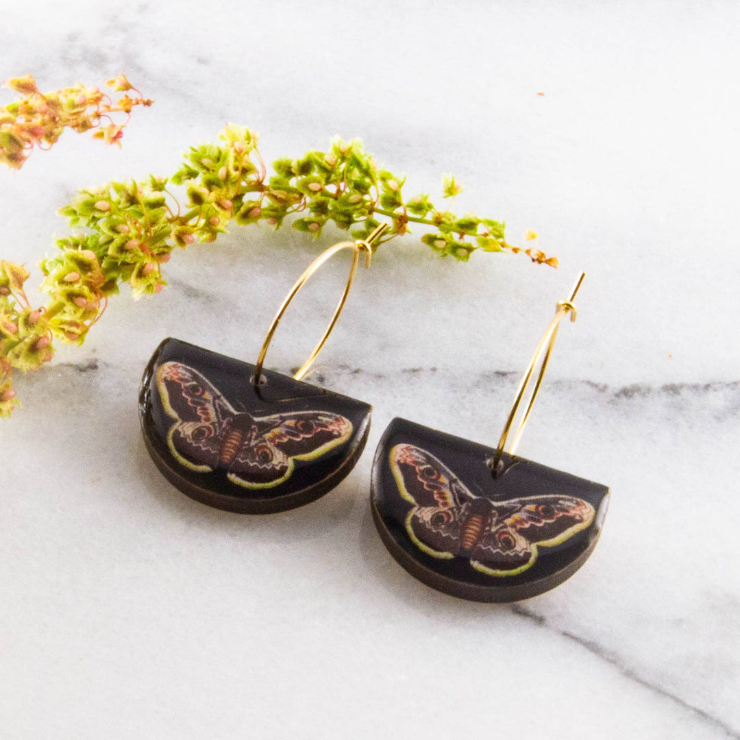 Moth-themed earrings on a marble surface with flowers.