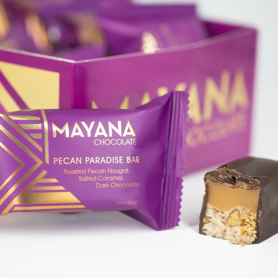 Mayana Chocolates | Stacy K Floral | A purple package with gold accents. Pecan Paradise Bar, toasted pecan nougat, salted caramel, dark chocolate. 1.5 oz