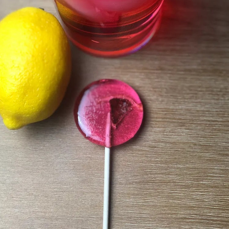 Good Lolli Lollipops | Pink Lemonade | A transparent pink lolli with a piece of lemon inside. Photo taken with a lemon and glass of pink lemonade in the background.