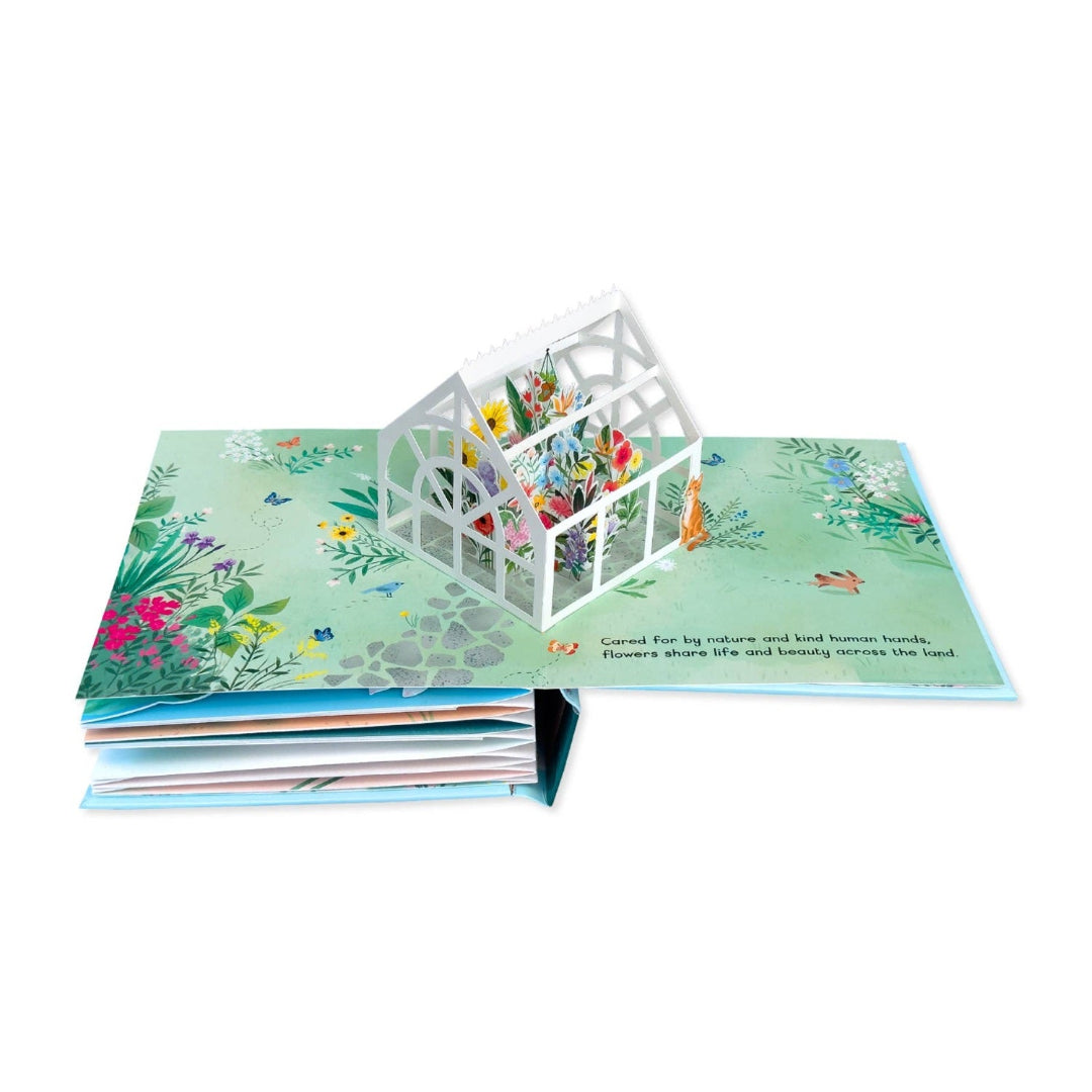 An open illustrated pop-up book showing a white greenhouse structure rising from a floral green cover, photographed on a clean white background.