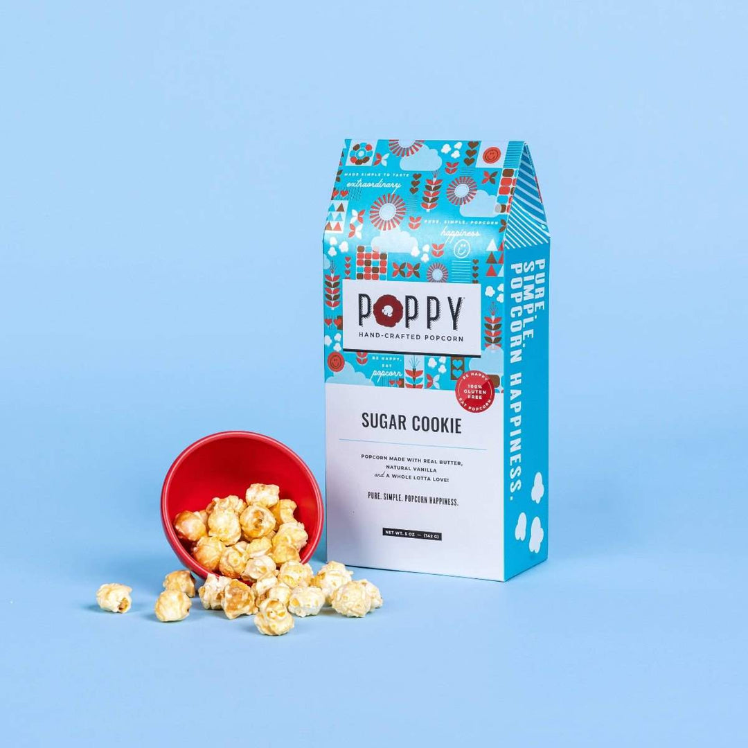 Poppy Hand-Crafted Popcorn | Sugar Cookie Gift Box Popcorn | A blue and white box with red accents. A small bowl of popcorn lays spilling kernels next to the box.