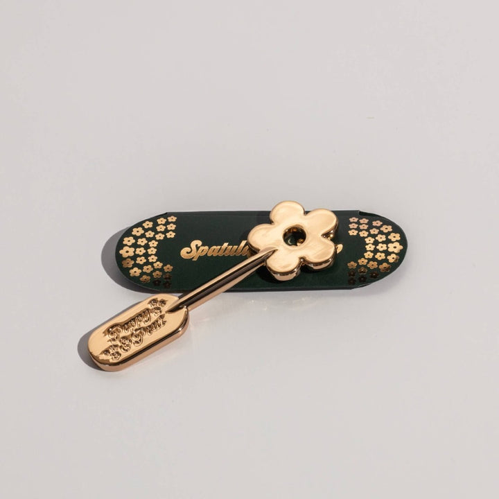 Poppy & Pout | Lip Care Spatula, Gold Daisy | A small gold spatula with a daisy on one side. The packaging is green with gold daisy accents.