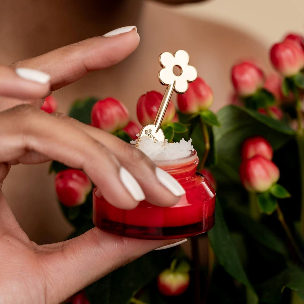 Poppy & Pout | Lip Care Spatula, Gold Daisy | A red lip scrub container with a gold daisy spatula. Being held by a model.