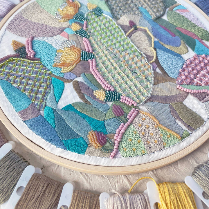 Embroidery hoop with colorful textile art and spools of thread on a wooden surface.