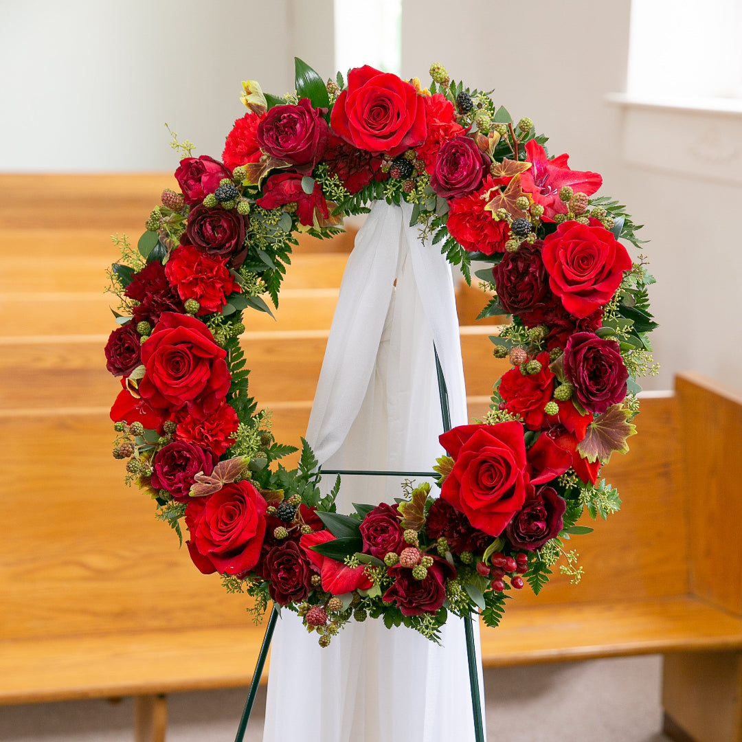 Floral wreath with red roses and greenery on a stand in a church setting.
