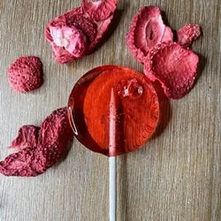 Good Lolli Lollipops | Ripe Strawberry | A transparent red lollipop with a piece of dried strawberry in the inside. Photo taken against a wood background with pieces of freeze dried strawberry.