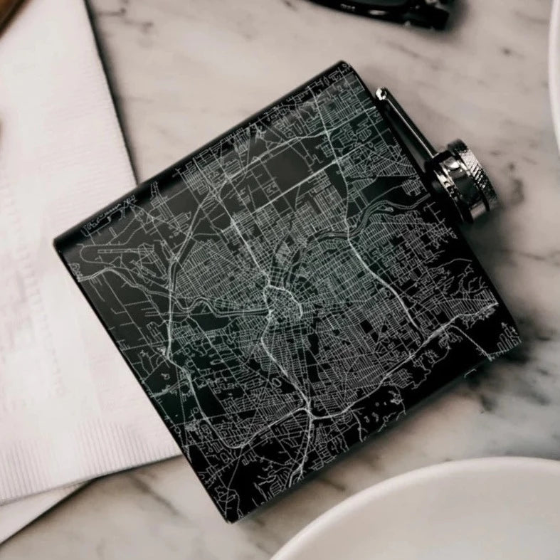 Rochester NY Engraved Flask | A black flask with an engraved silver map of Rochester NY. Flask is on its side against a marble background.