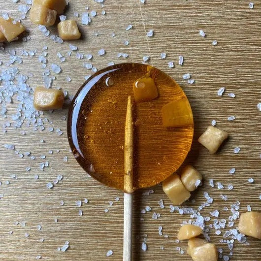 Good Lolli Sea Salted Caramel Lollipop | A caramel colored lollipop with pieces of caramel and salt. Photo taken against a wooden background with caramel and salt placed around.