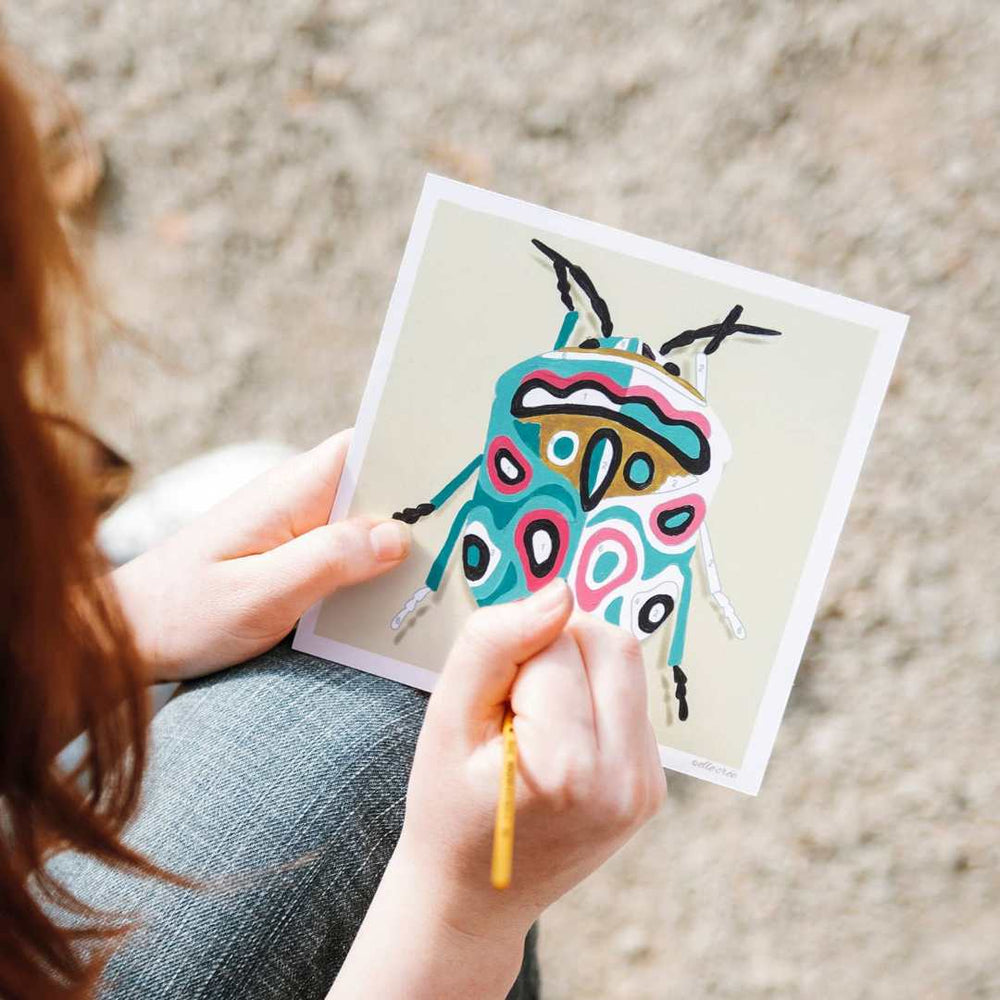 She Creates | Person holding a colorful beetle illustration on a card