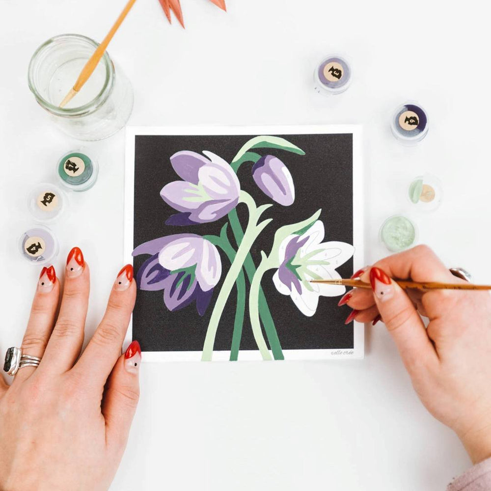 She Creates | Person painting a floral paint by number on a small canvas with art supplies around.