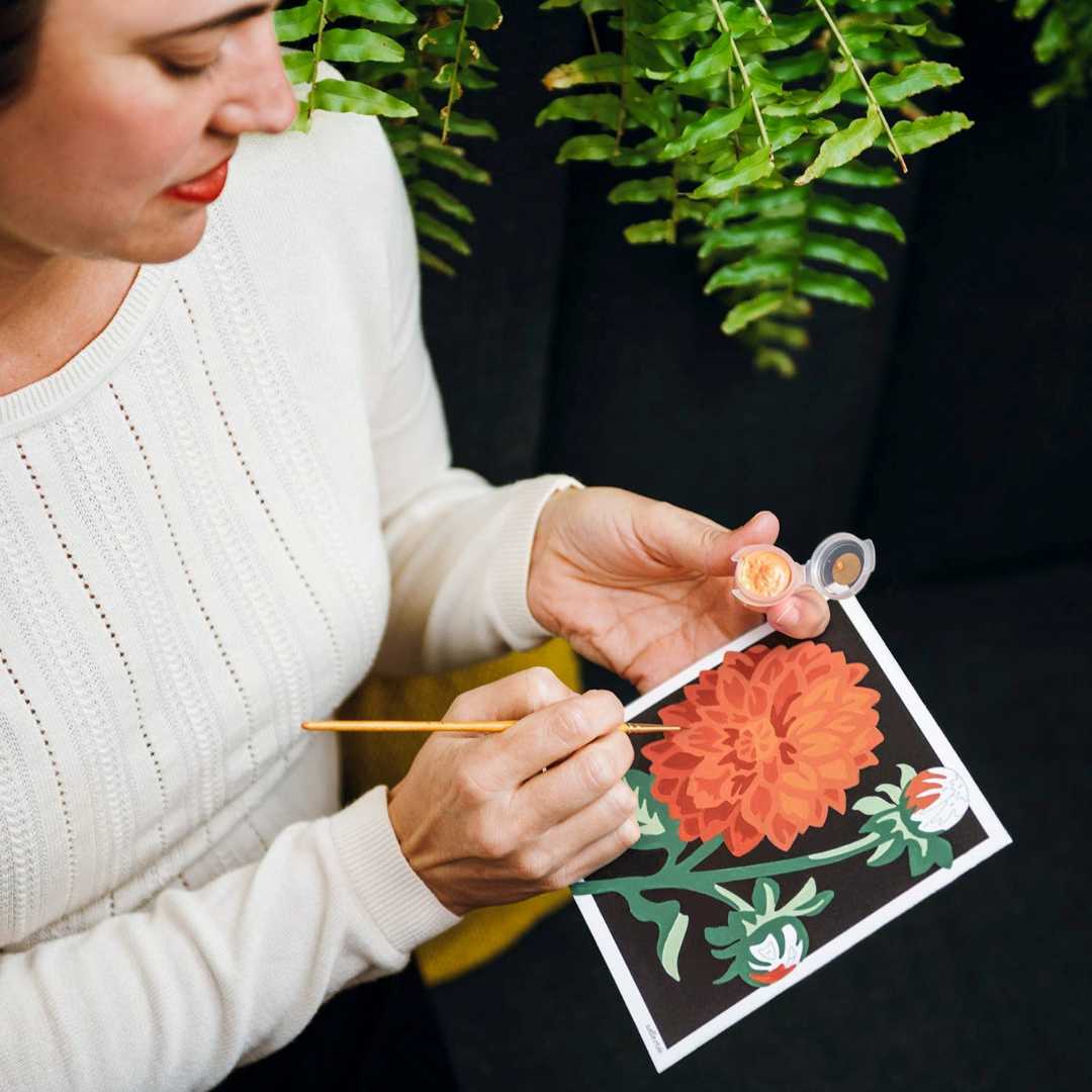 She Creates | Woman painting a floral design on a canvas with a plant in the background.
