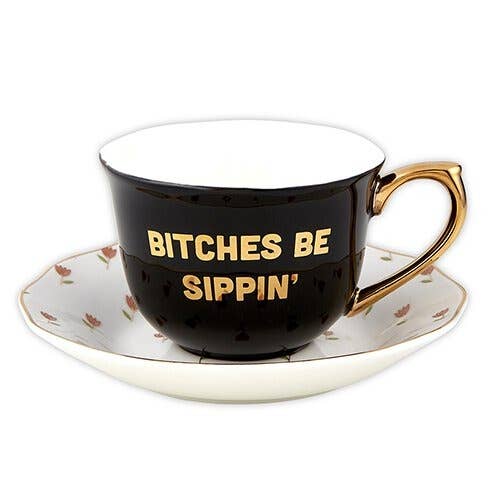 Slant | Teacup with text "Bitches Be Sippin' | A black teapot with gold accents and a white saucer with little pink flowers.